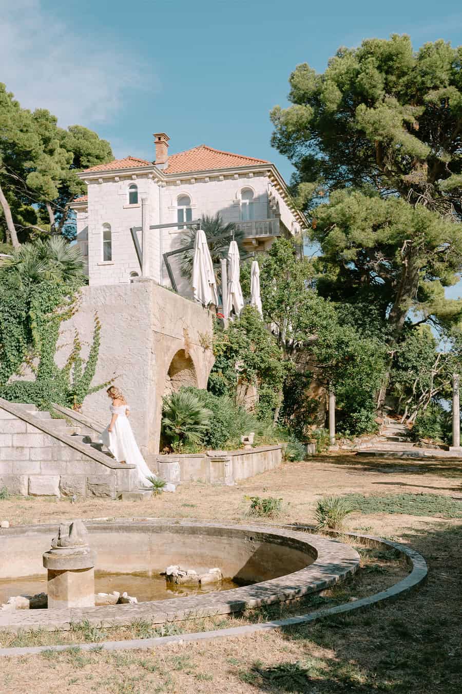 Široki kadar mladenke u vjenčanici koja se penje vanjskim kamenim stepenicama u mediteranskom vrtu ispred stare kamene vile i fontane u Dubrovniku.