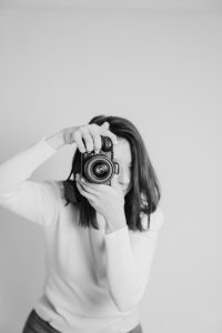 Crno-bijeli portret žene fotografkinje u studiju koja drži fotoaparat ispred lica u trenutku snimanja.