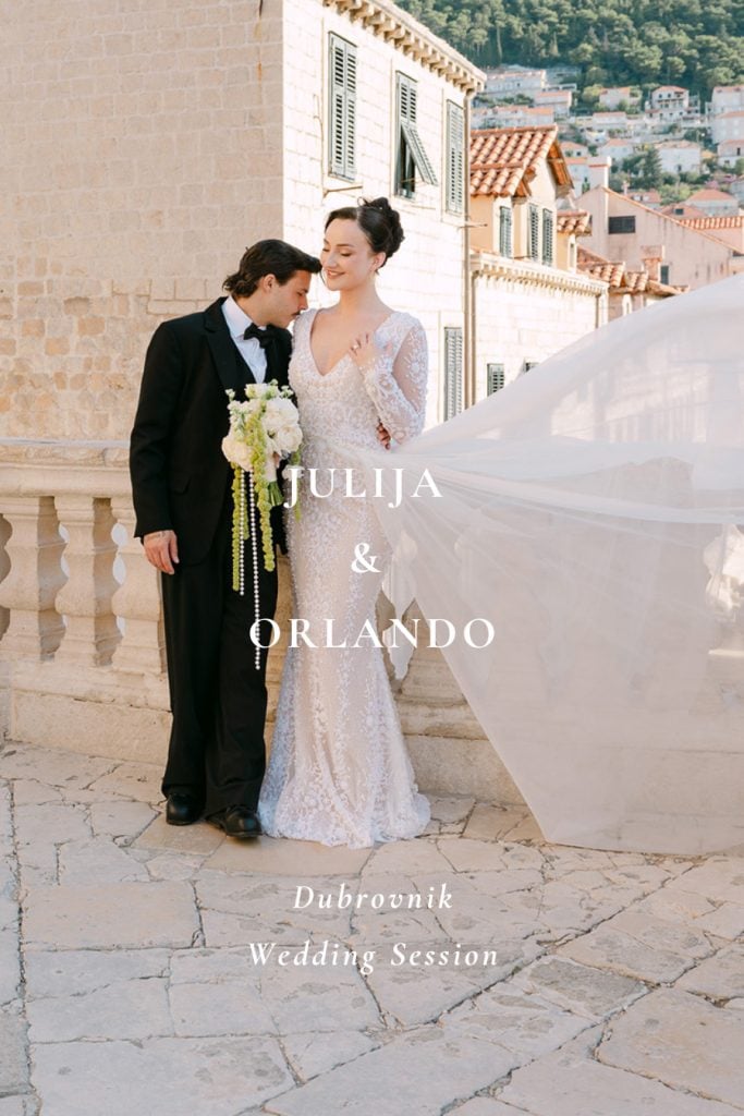 Mladoženja ljubi mladenku u rame dok njezin dugi veo leti na kamenoj terasi sa starim gradom u pozadini tijekom svadbenog sessiona u Dubrovniku.