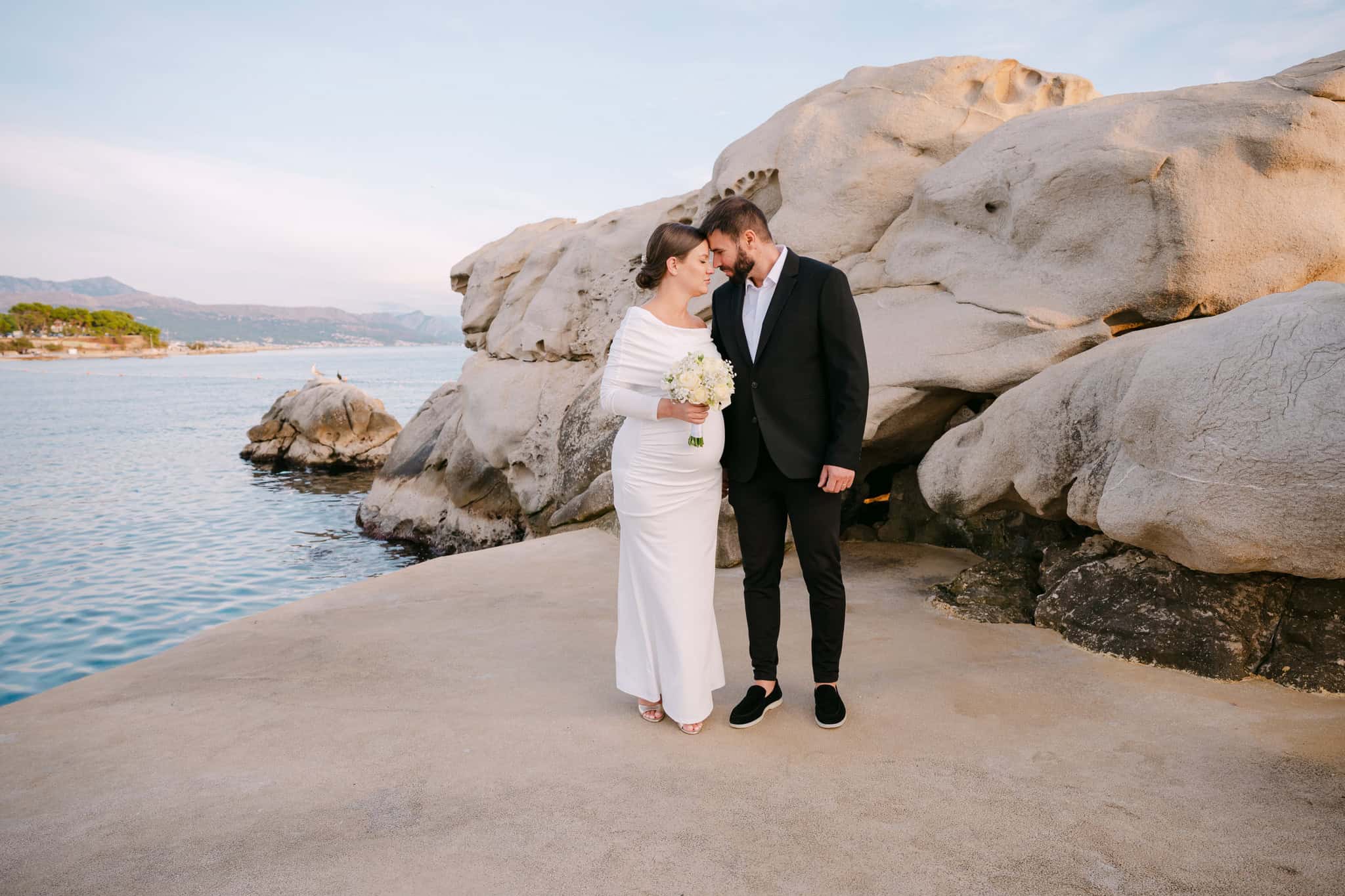 Svadbeni par stoji blizu plaže s velikim stijenama i morem u pozadini.