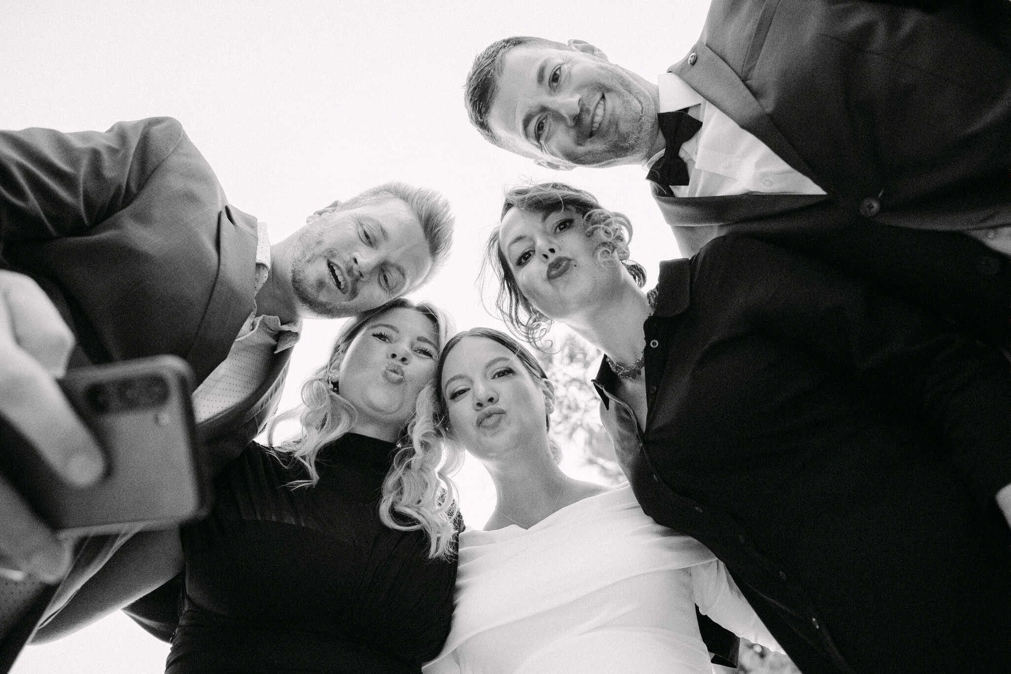 Grupa prijatelja na vjenčanju, formalno odjeveni, nasmijani i snimaju selfie.