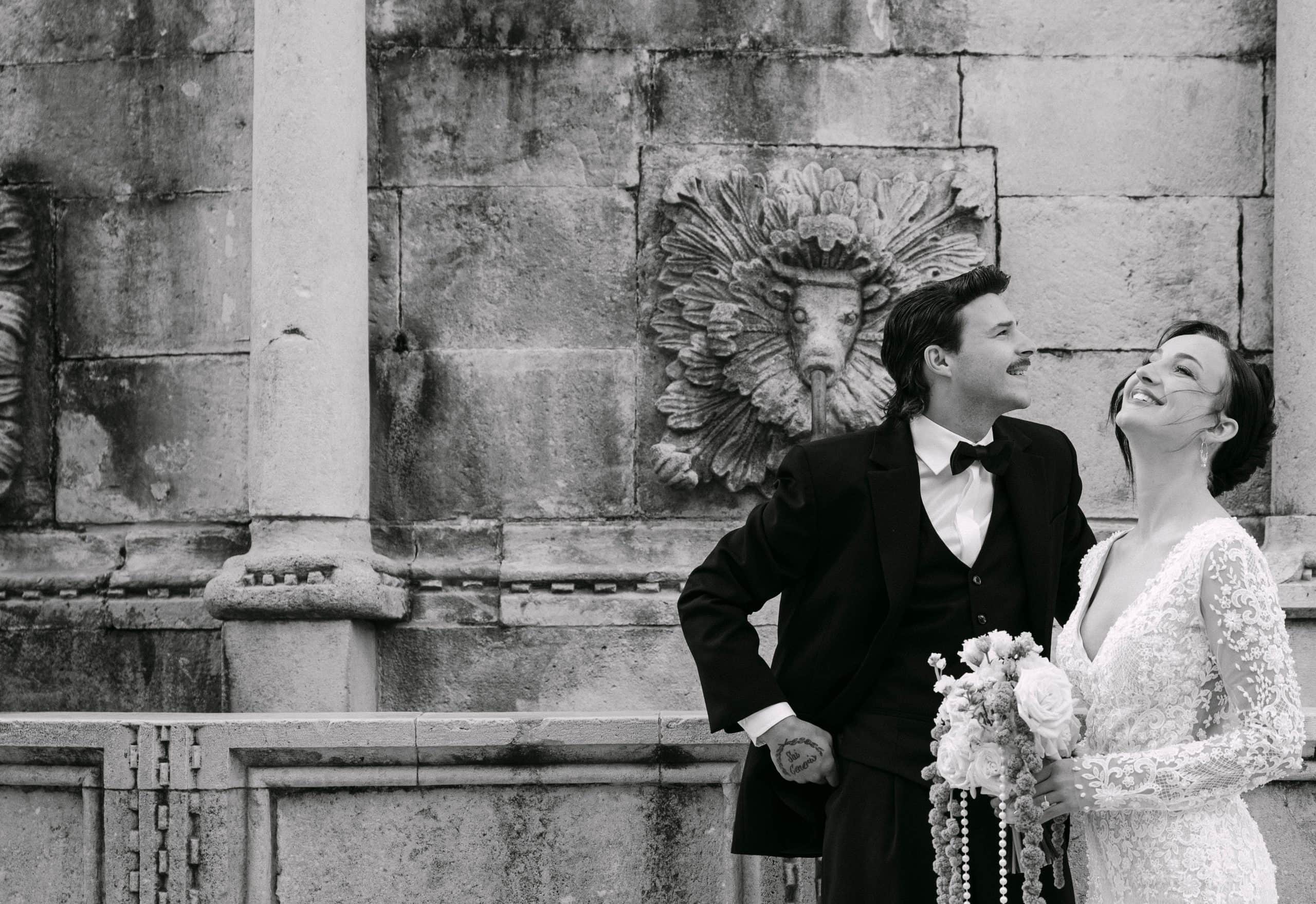 Dubrovnik wedding fotograf - dokumentarni crno-bijeli kadar; mladenka se radosno smije dok stoji uz mladoženju ispred teksture starog dubrovačkog kamena.