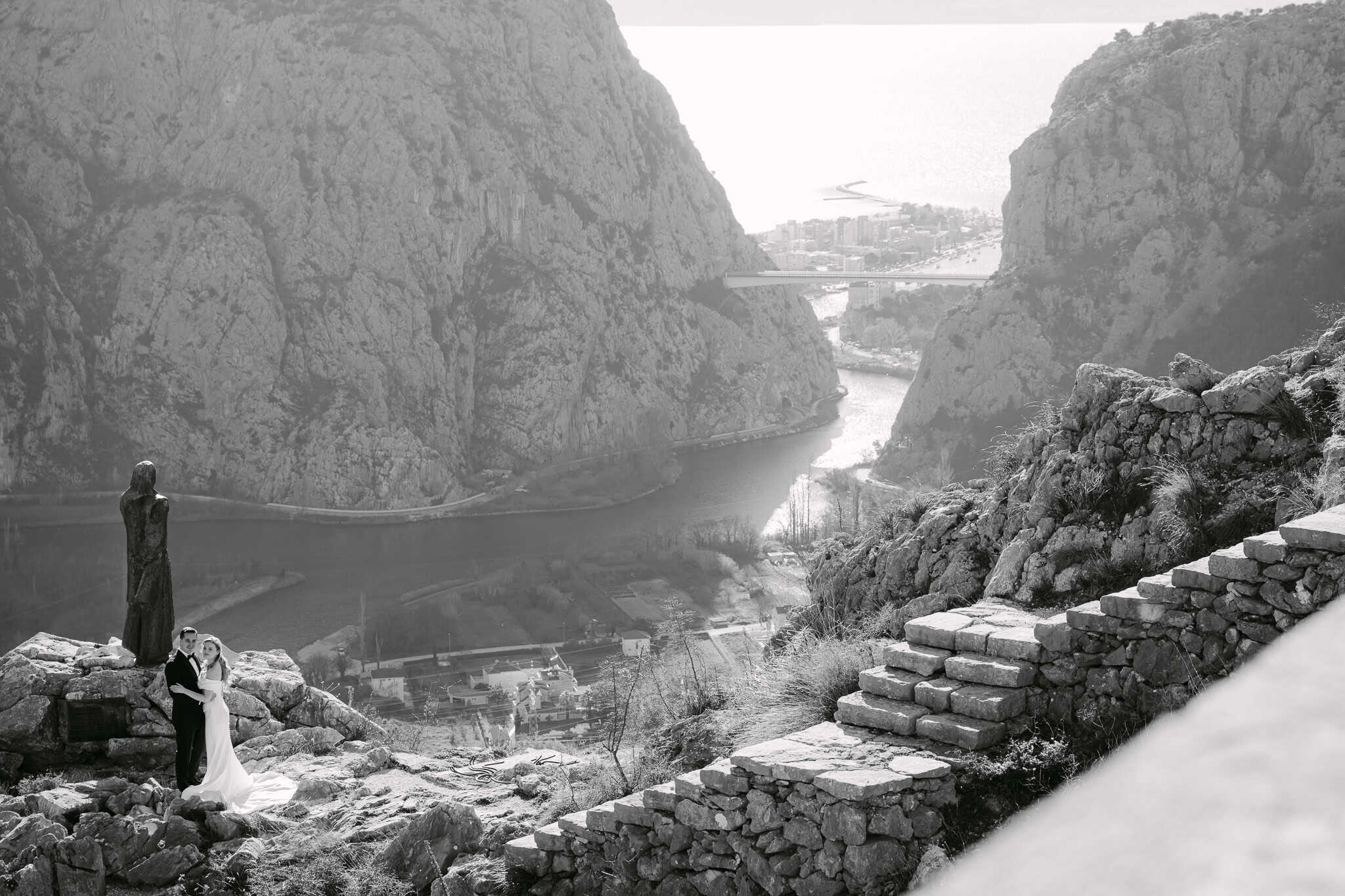 Crno-bijela fotografija svadbenog para na vidikovcu s pogledom na ušće rijeke Cetine i grad Omiš.