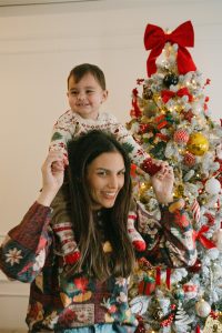 Blagdansko obiteljsko fotografiranje u domu gdje majka drži malo dijete na ramenima ispred ukrašenog božićnog drvca.