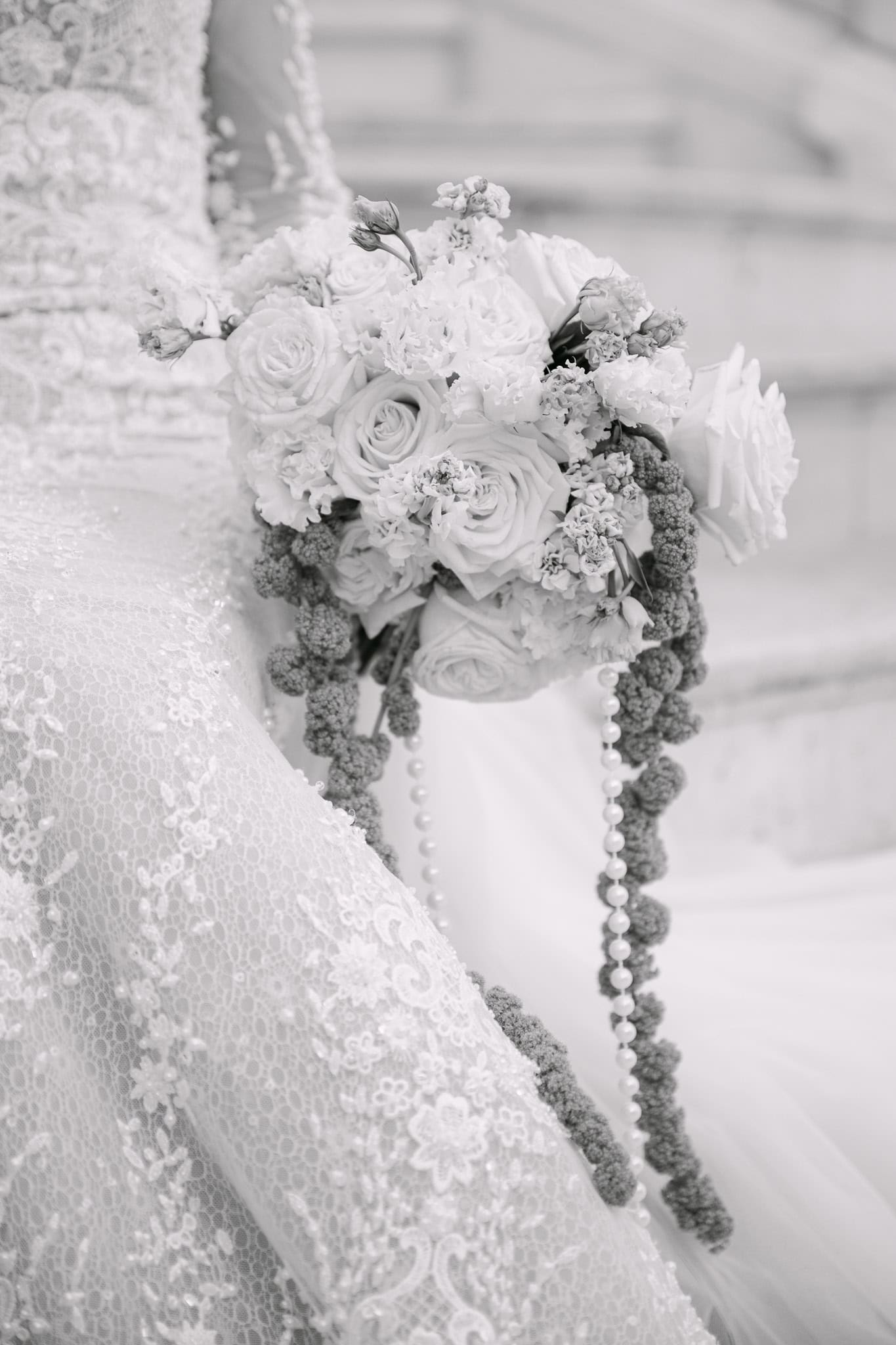 Krupni plan raskošnog vjenčanog buketa ukrašenog dugačkim nizom bisera; umjetnički crno-bijeli detalj koji naglašava teksturu čipkaste haljine.