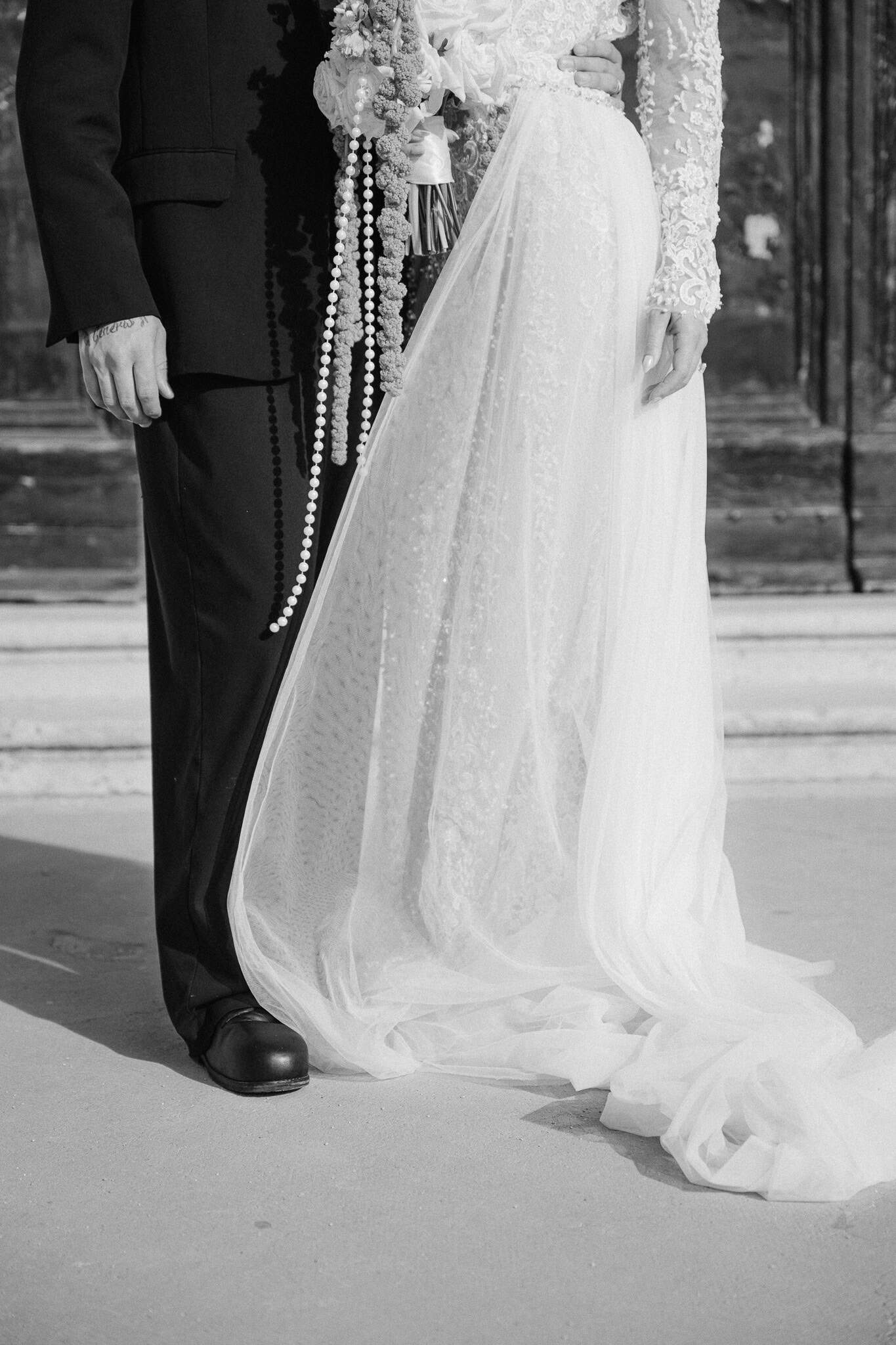 Dubrovnik destination wedding – umjetnički crno-bijeli detalj vjenčanja; krupni plan elegantnog dugog šlepa na haljini i padajućih bisernih ukrasa na sunčevoj svjetlosti ispred starogradske arhitekture.