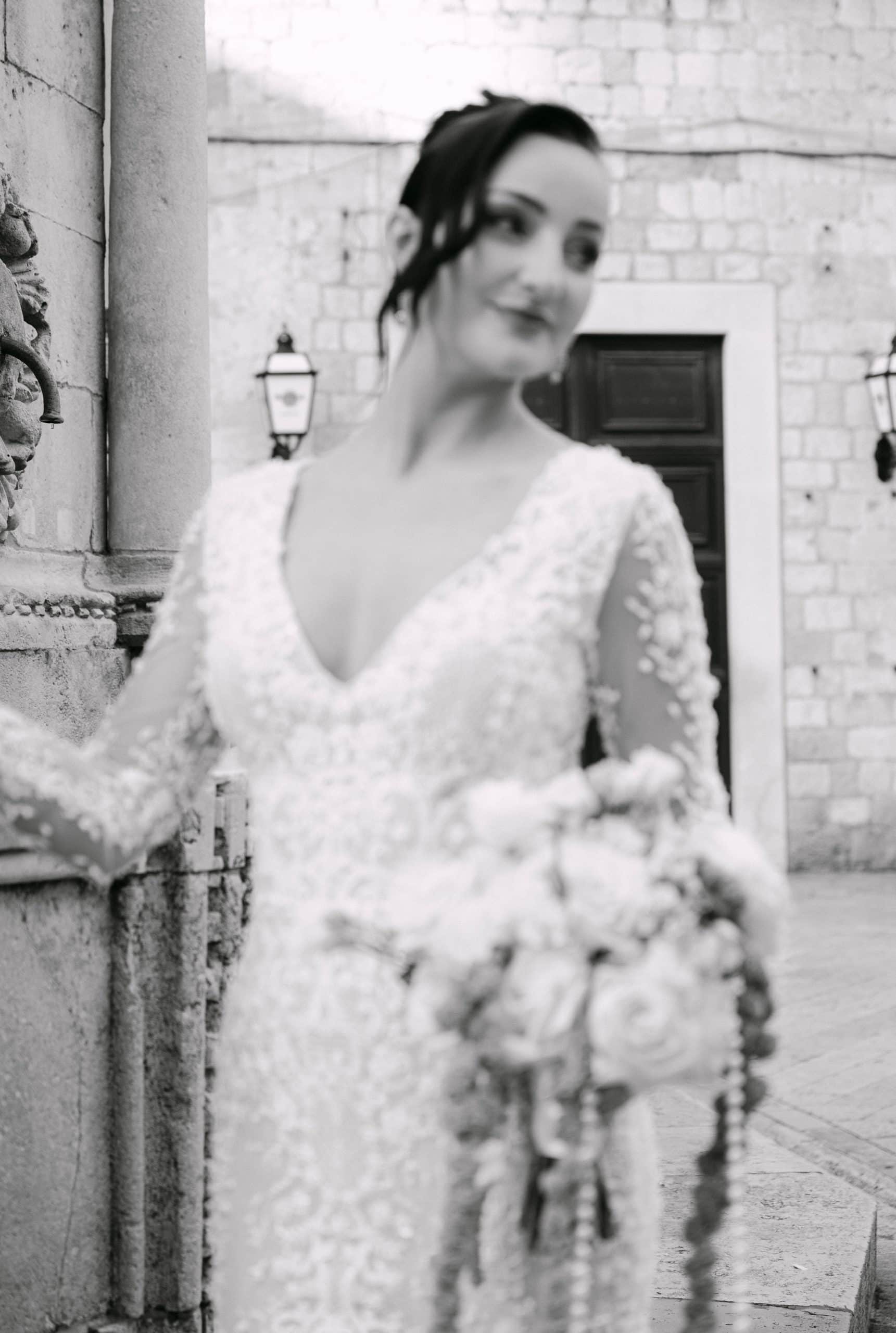 Vjenčanje u Dubrovniku - elegantni crno-bijeli portret mladenke; nevjesta u čipkastoj haljini s bogatim bidermajerom ispred starih dubrovačkih kamenih vrata.