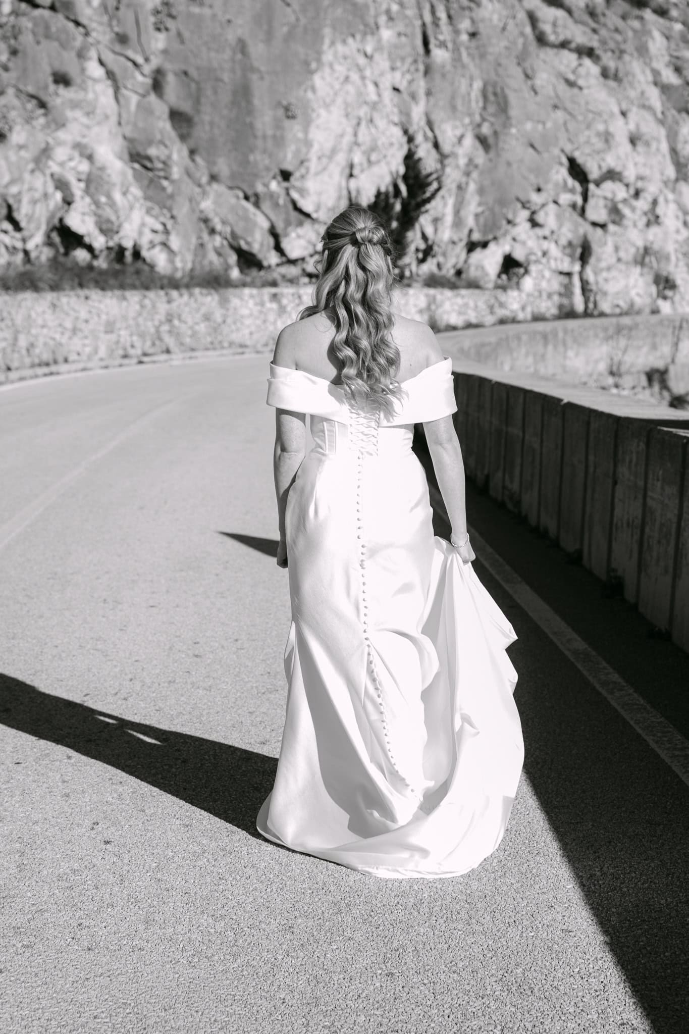 Crno-bijela fotografija mladenke u dugoj vjenčanici snimljene s leđa kako hoda asfaltiranom cestom uz stijene.