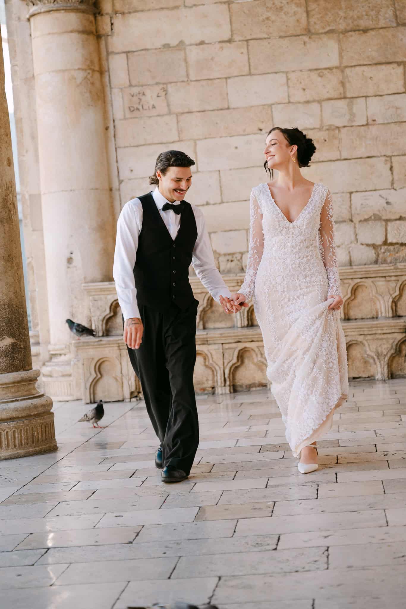 Vjenčanje u Dubrovniku – spontani kadar; mladoženja u prsluku i mladenka u čipkastoj vjenčanici nasmijano šetaju kroz dubrovačke zidine uz golubove.
