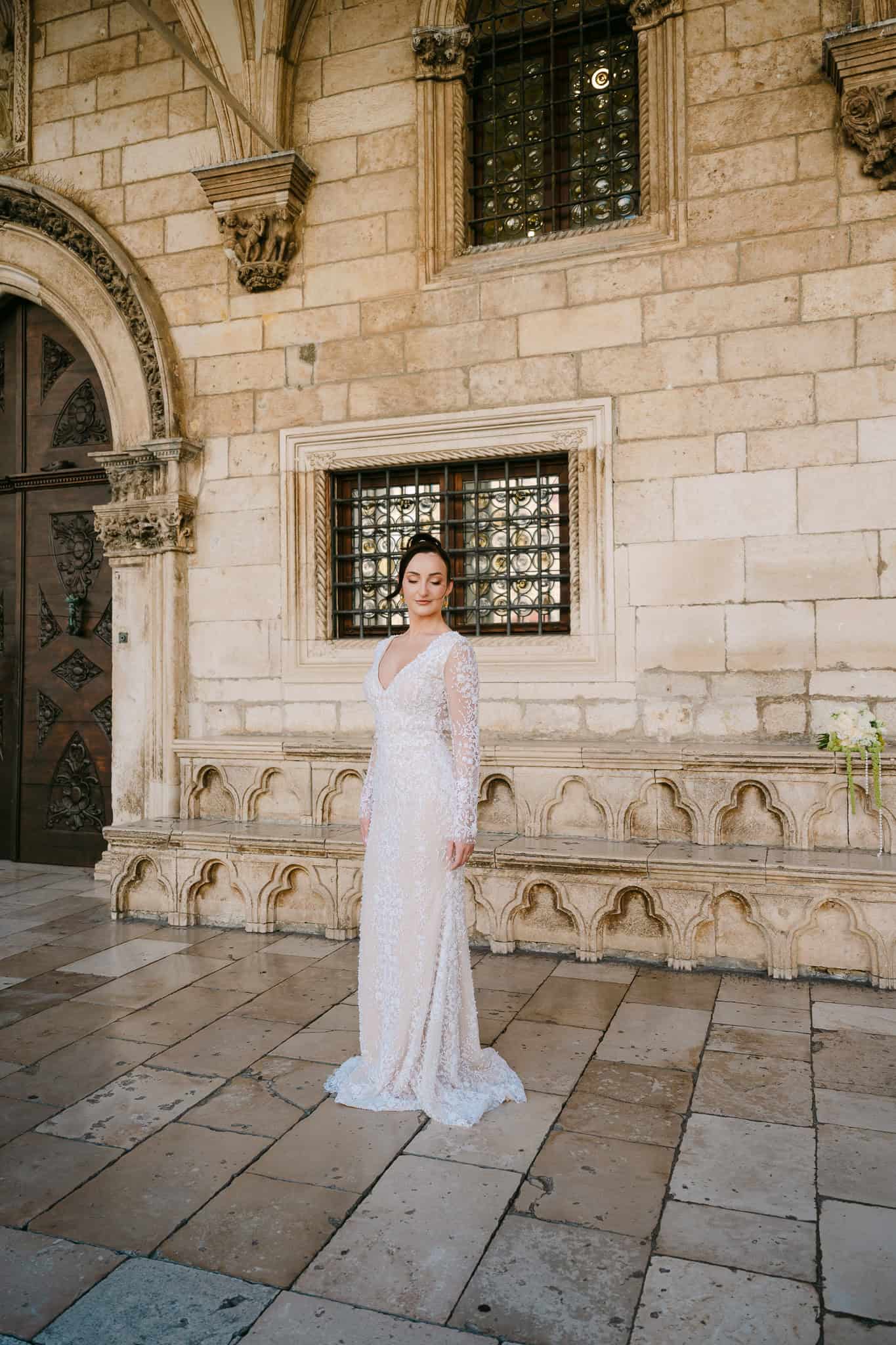 Vjenčanje Dubrovnik - široki portret mladenke u punoj visini; nevjesta u dugoj čipkastoj haljini pozira ispred arhitekture Starog grada i kamenih prozora.