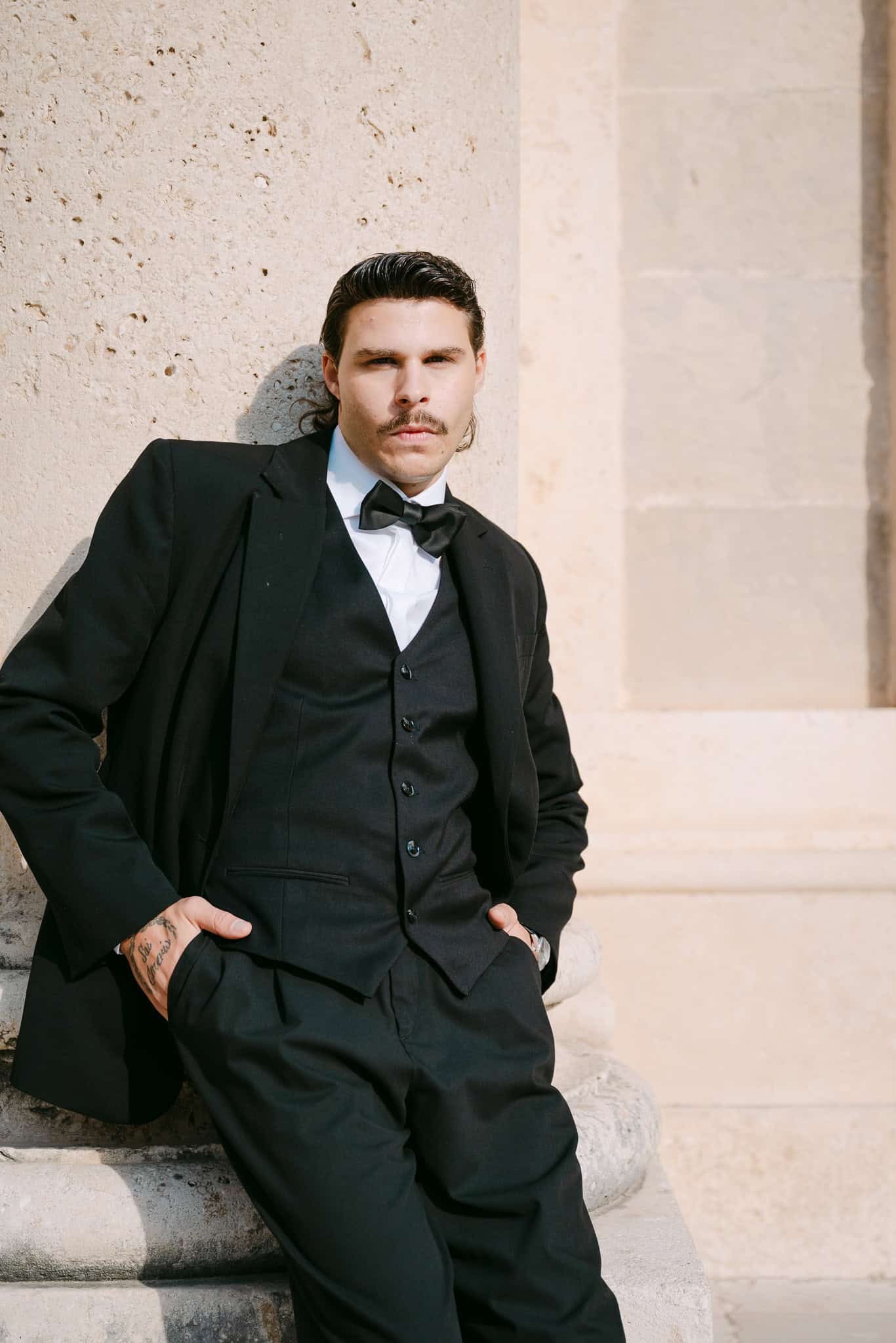 Dubrovnik elopement – klasični portret mladoženje na otvorenom; elegantan mladoženja u crnom odijelu opušteno pozira naslonjen na osunčani povijesni kameni stup.