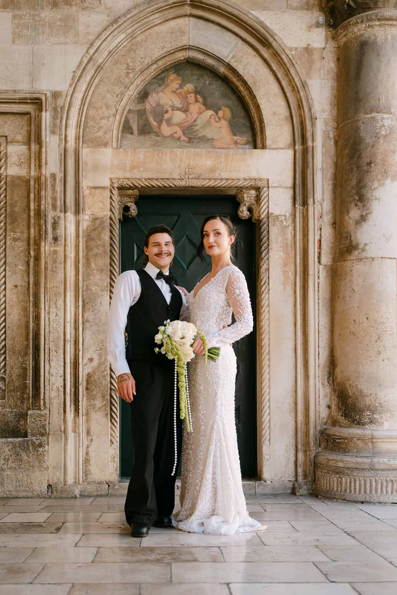 Dubrovnik, povijesna jezgra - klasični vjenčani portret; elegantni mladenci poziraju gledajući u kameru ispred raskošnog kamenog ulaza s freskom.