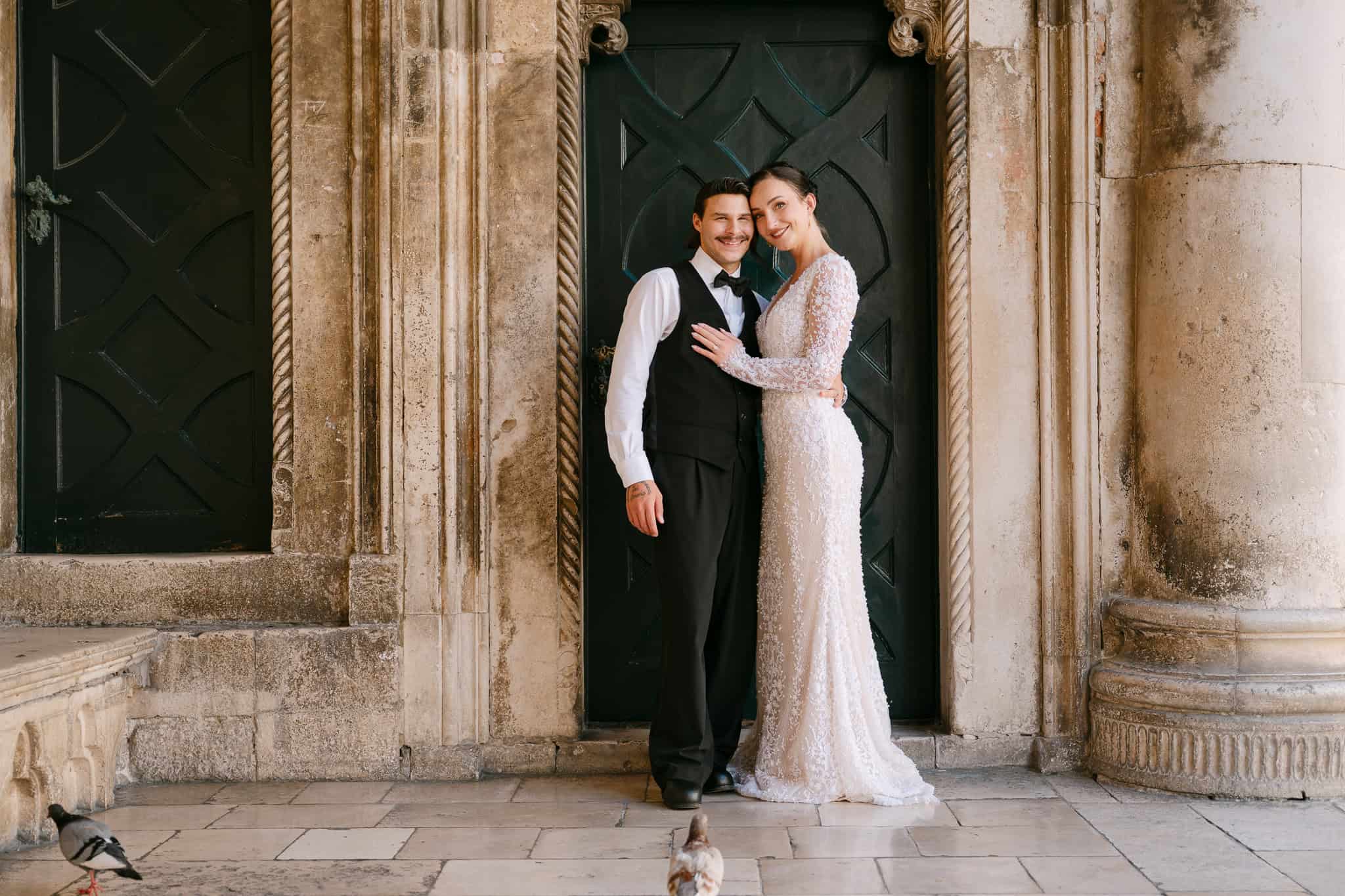 Klasični vjenčani portret; nasmiješeni par pozira ispred povijesnih masivnih vrata i fasade s freskom, s golubovima na kamenom pločniku.