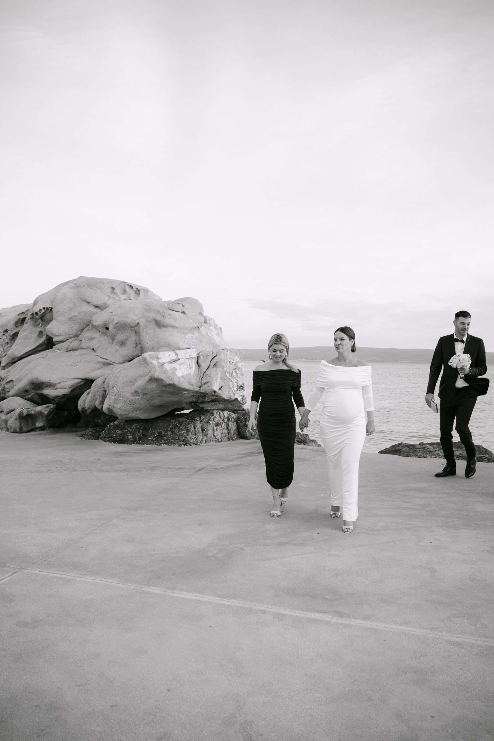 Crno-bijeli prikaz mladenke i kume na plaži u Splitu; vjenčani session uz more dok kum u pozadini drži buket i torbicu.