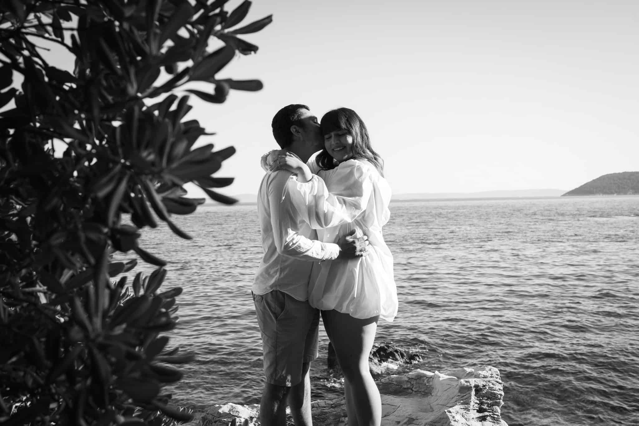 Zaručeni par na obali mora, snimljen u crno-bijeloj boji, uživa u romantičnom trenutku uz more.