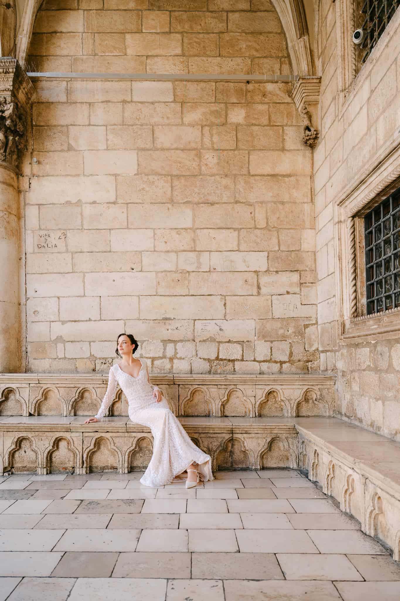 Dubrovnik wedding session – široki portret mladenke u boji; elegantna nevjesta odmara na kamenoj klupi okružena impresivnom arhitekturom Starog grada.