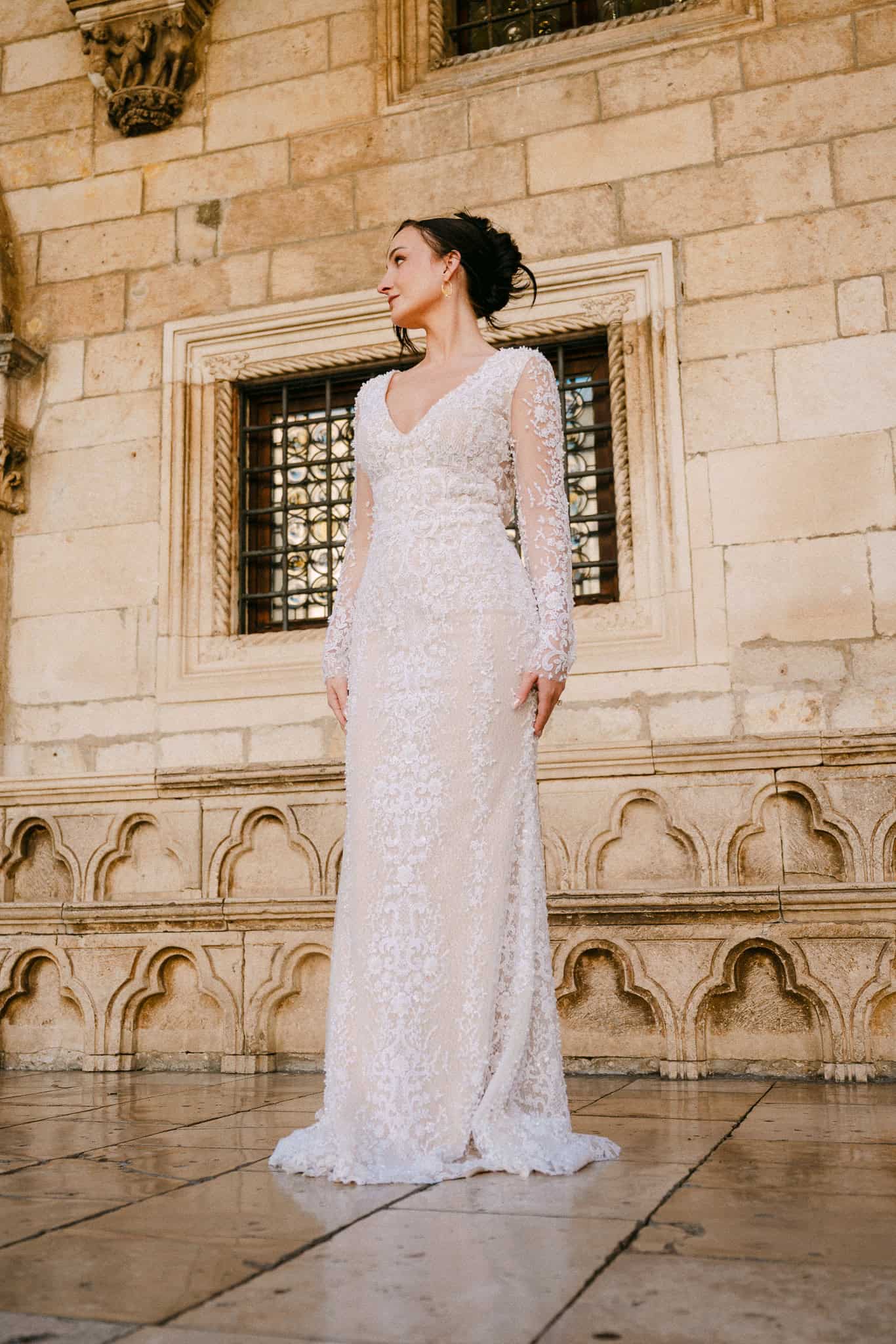 Stari grad Dubrovnik – klasičan portret mladenke; nevjesta u bijeloj haljini dugih rukava gleda u stranu pozirajući na uglačanom pločniku ispred povijesnih zidina.