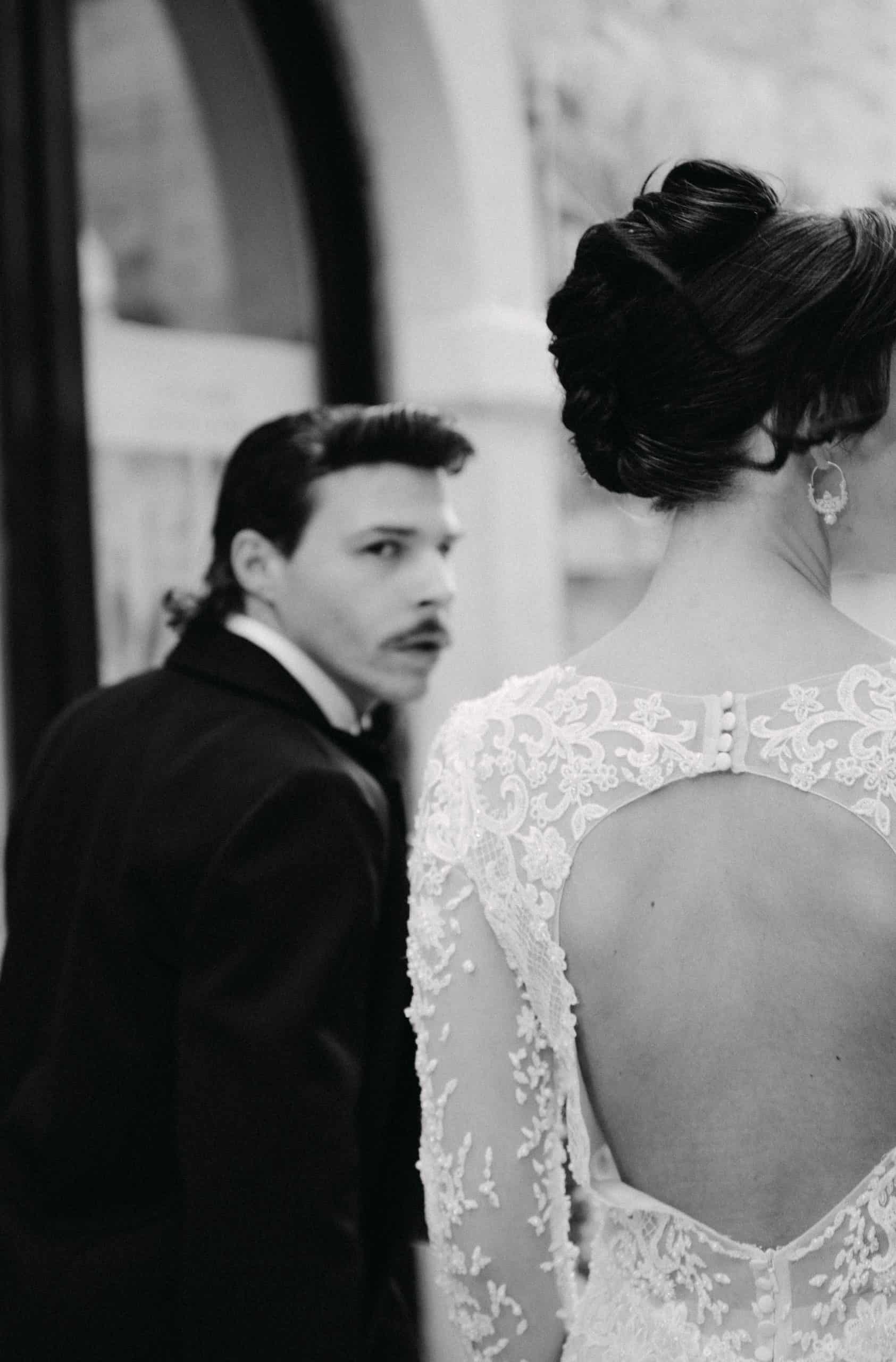 Vjenčani par na fotografiji, fokus na žensku haljinu s čipkom i mladoženja u pozadini.