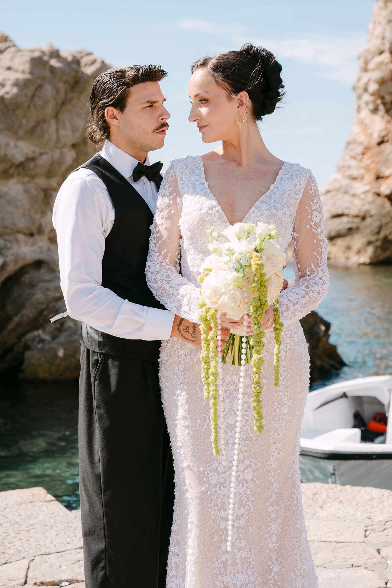Dubrovnik vjenčani fotograf - romantični pogled mladenaca; mladoženja u crnom prsluku i mladenka u čipkastoj haljini uživaju na osunčanom pristaništu.