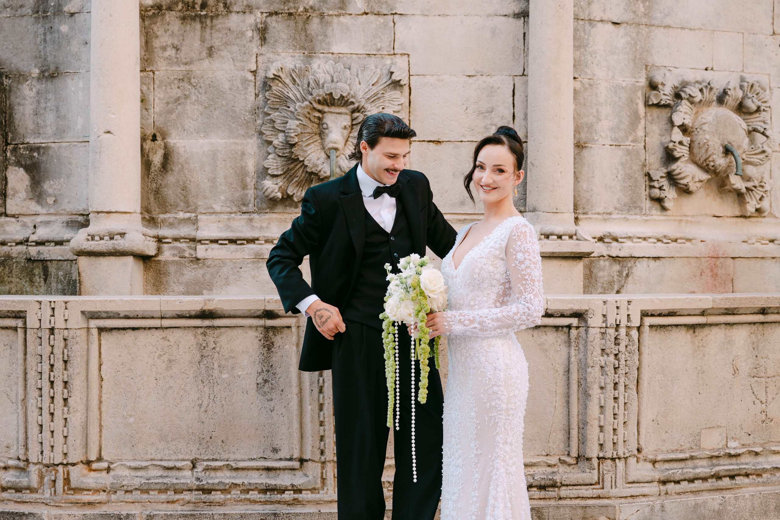 Vjenčanje u Dubrovniku - opušteni portret mladenaca ispred stare kamene fontane; nasmiješeni par u elegantnom izdanju na sessionu na otvorenom.