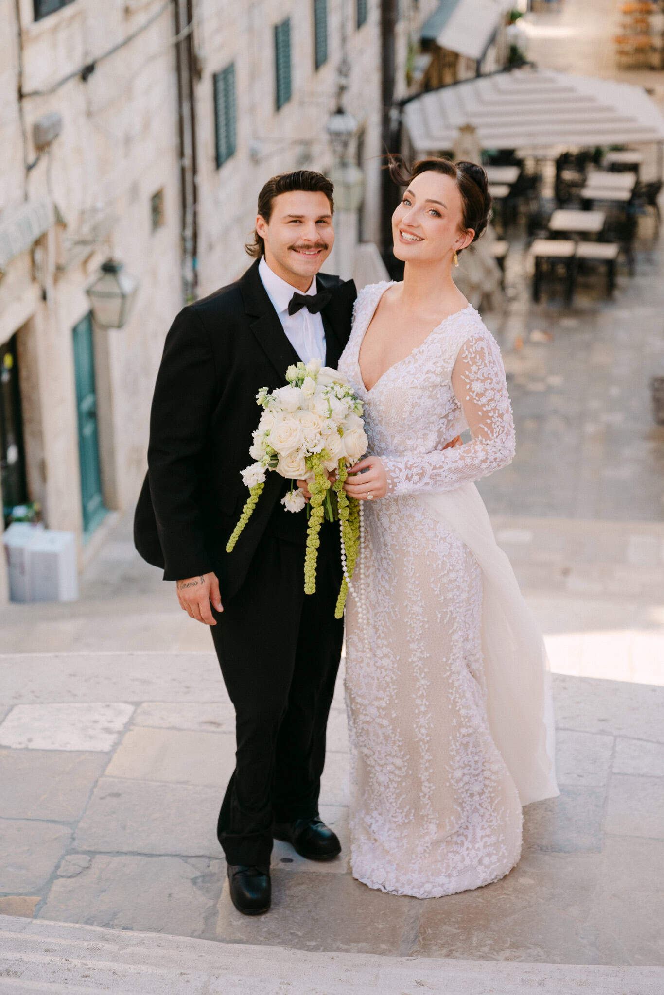 Opušteni pozerski trenutak na suncem okupanim ulicama; mladenci stoje na kamenim stepenicama Starog grada, mladenka drži svijetli bidermajer s padajućim zelenilom dok se oboje blago osmjehuju prema kameri.