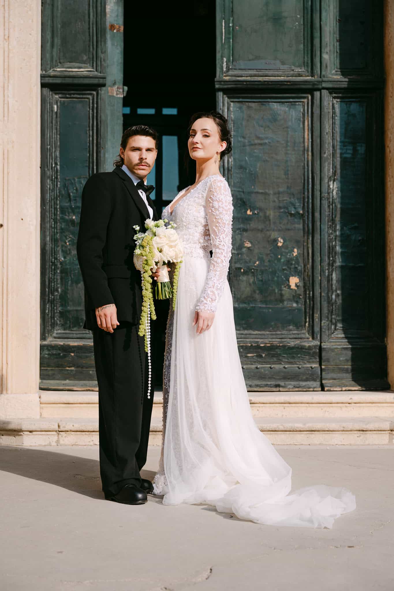 Vjenčanje u starom gradu Dubrovniku – elegantni lifestyle portret mladenaca na otvorenom; par pozira u punoj visini ispred masivnih, starinskih drvenih vrata s izraženom teksturom i povijesnim šarmom.
