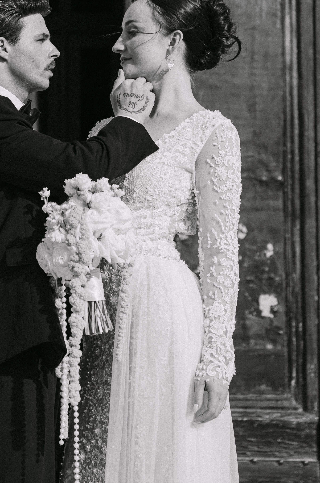 Vjenčani fotograf Dubrovnik na otvorenom - intimni crno-bijeli portret; mladoženja nježno dodiruje lice nasmiješene nevjeste s naglaskom na raskošan kaskadni vjenčani buket s biserima.
