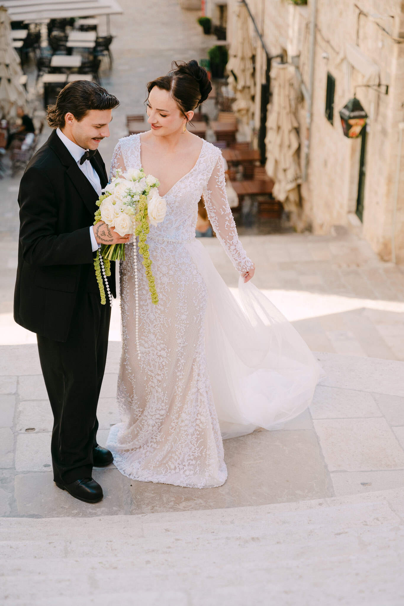 Opuštena šetnja mladenaca kamenim stubama Dubrovnika; nevjesta u elegantnoj haljini s bogatim bidermajerom i mladoženja u crnom odijelu uživaju u zajedničkom trenutku na otvorenom ispred vizure gradskih ulica.