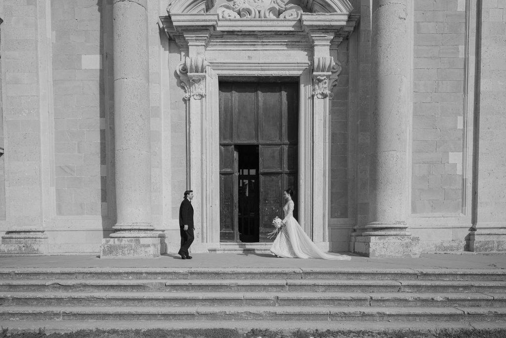 Luksuzno vjenčanje Dubrovnik – široki crno-bijeli kadar; mladenci u elegantnom editorijal stilu poziraju na suprotnim stranama ispred monumentalnog kamenog ulaza sa stupovima tijekom outdoor sessiona.