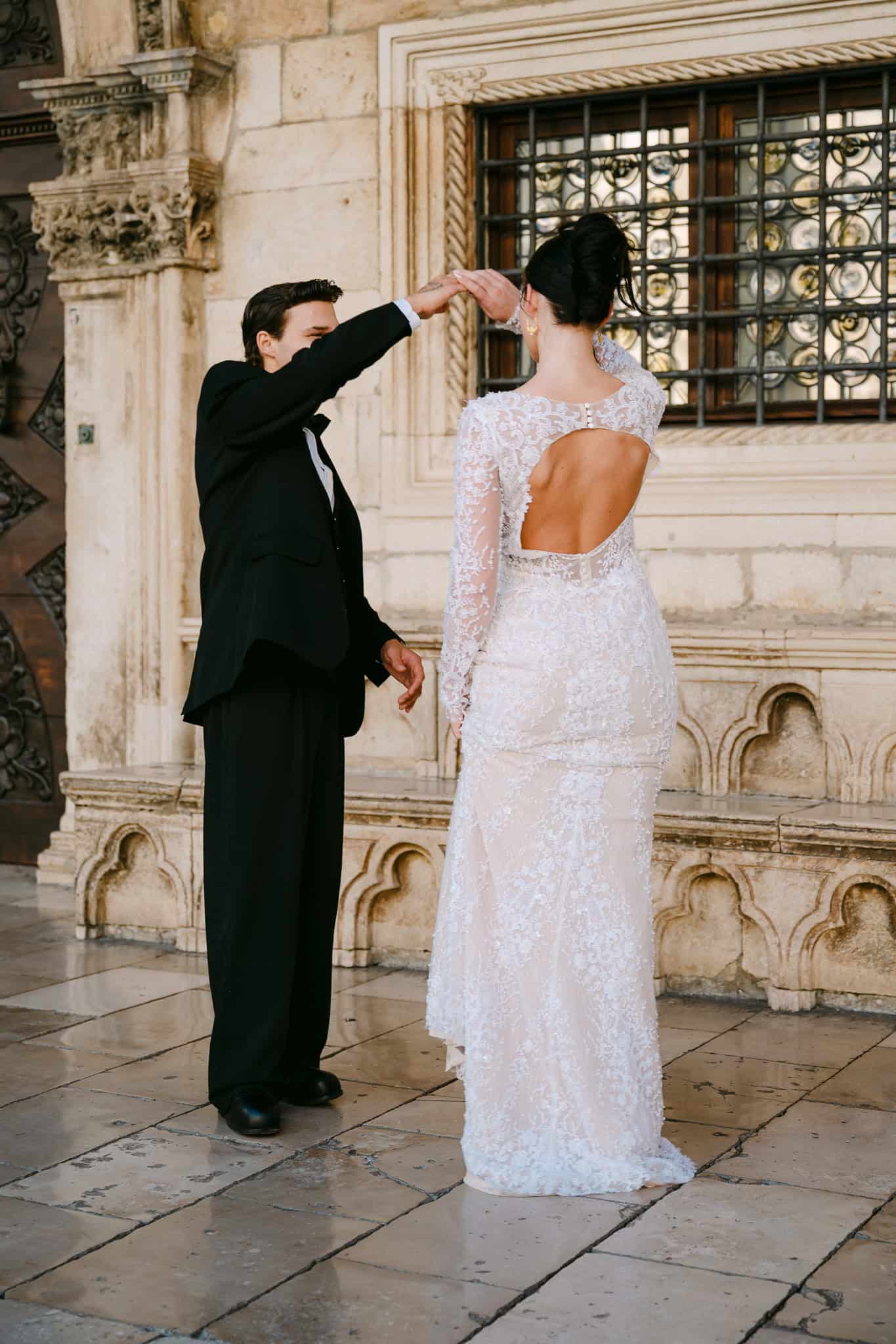 Romantičan ples mladenaca; mladoženja okreće nevjestu u elegantnoj čipkastoj vjenčanici s otvorenim leđima ispred povijesne kamene arhitekture i ukrašenih prozora u starom dijelu grada Dubrovnika.