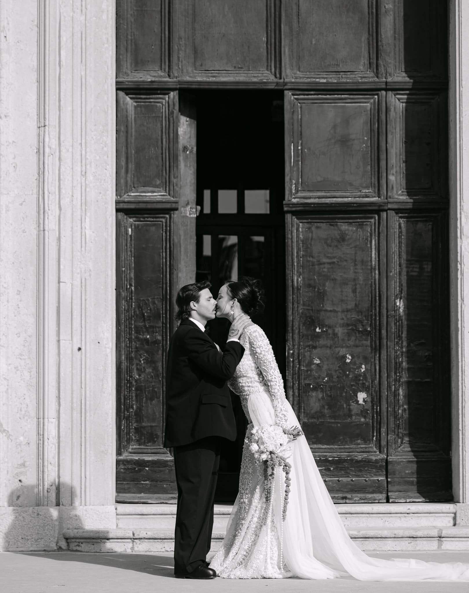 Luksuzno vjenčanje u Dubrovniku – dramatičan crno-bijeli kadar poljupca na otvorenom; mladenci u romantičnom trenutku stoje ispred impresivnih, poluotvorenih monumentalnih vrata koja naglašavaju bogatu povijest grada.