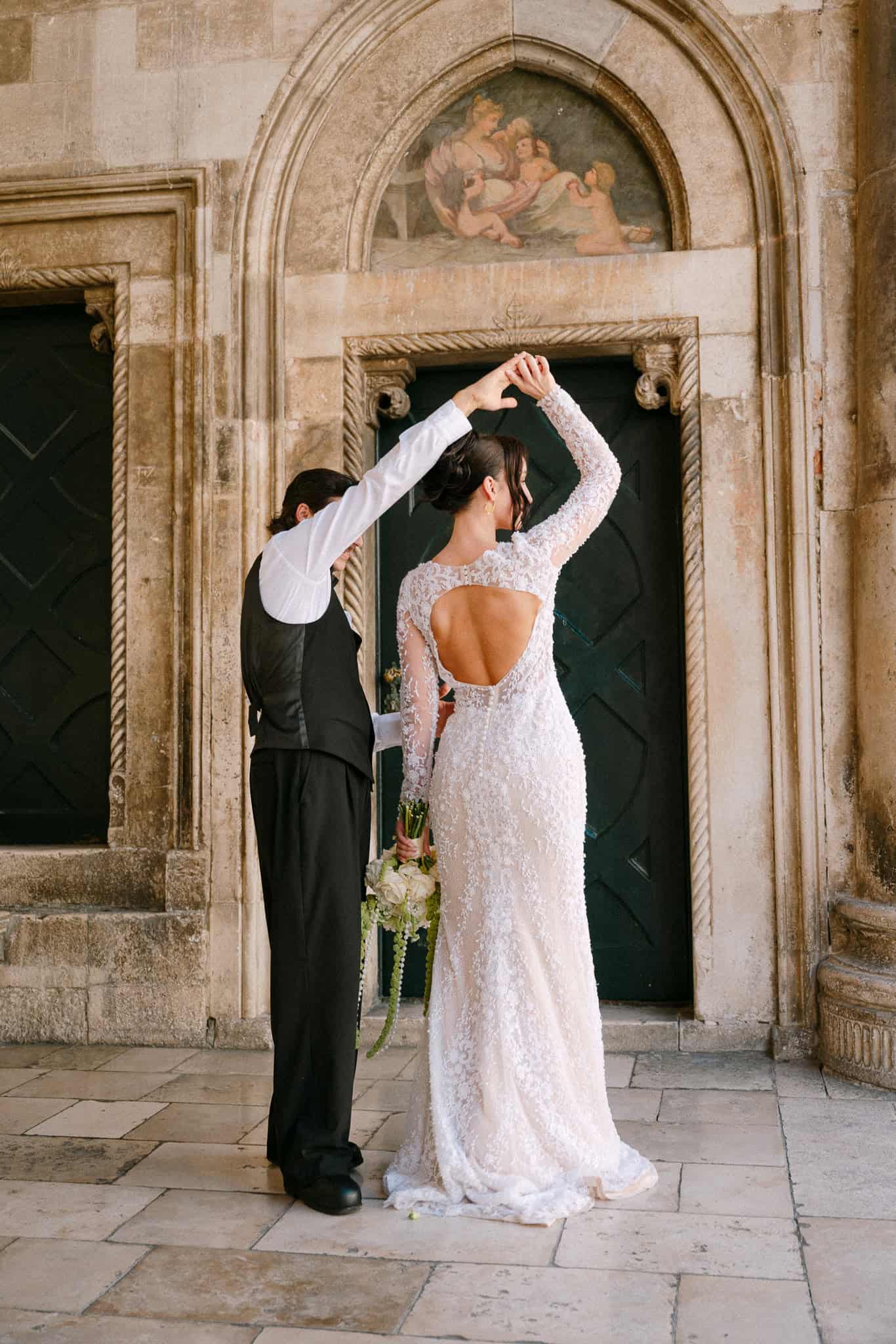 Dubrovnik - romantični ples mladenaca; mladoženja okreće mladenku u vjenčanici s otvorenim leđima ispred povijesnih kamenih zelenih vrata s freskom.