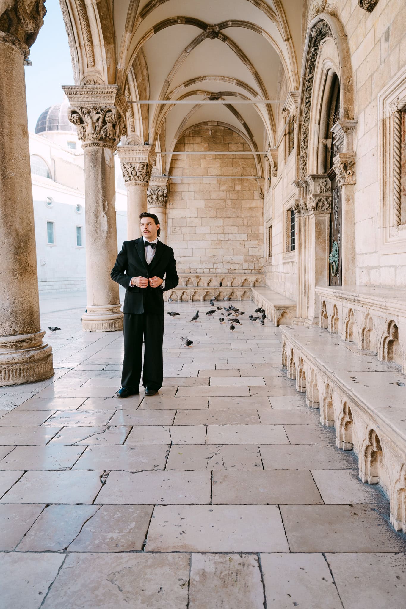 Portret mladoženje na otvorenom; mladoženja u elegantnom crnom odijelu pozira ispod povijesnih dubrovačkih arkada.