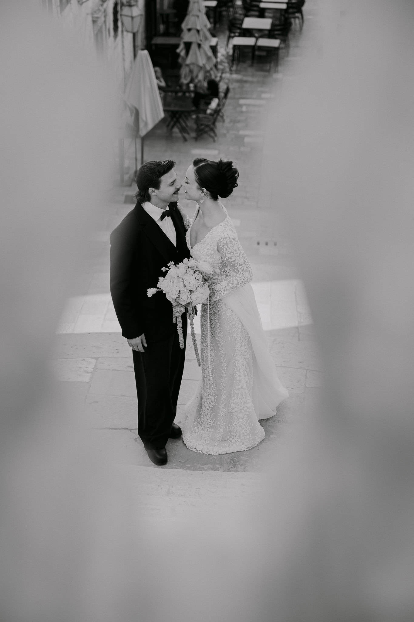 Filmski crno-bijeli portret mladenaca na otvorenom; par se nježno dodiruje nosovima u romantičnom trenutku, dok zamućeni okvir u prvom planu stvara intimnu atmosferu usred kamenih ulica Dubrovnika.