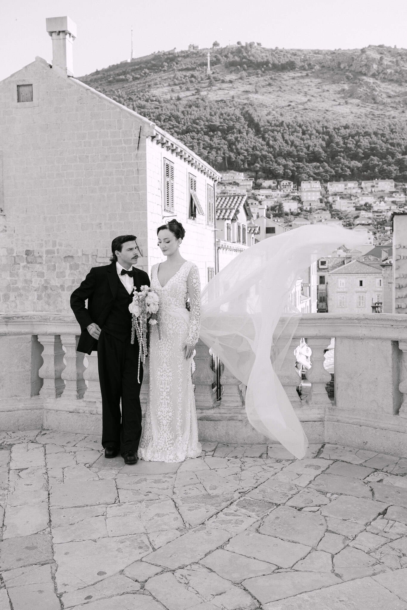 Dugački prozirni veo mladenke vijori na vjetru ispred krovova starog grada Dubrovnika i brda Srđ u pozadini dok par pozira na kamenoj terasi.