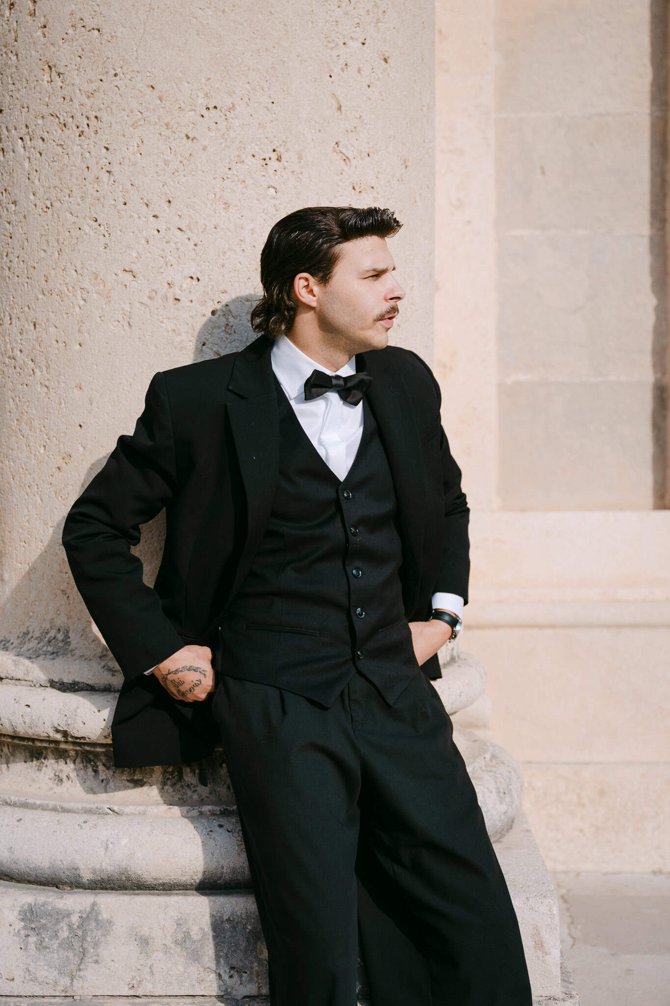 Mladoženja u crnom odijelu naslonjen na masivni kameni stup monumentalnih arkada grada Dubrovnika.