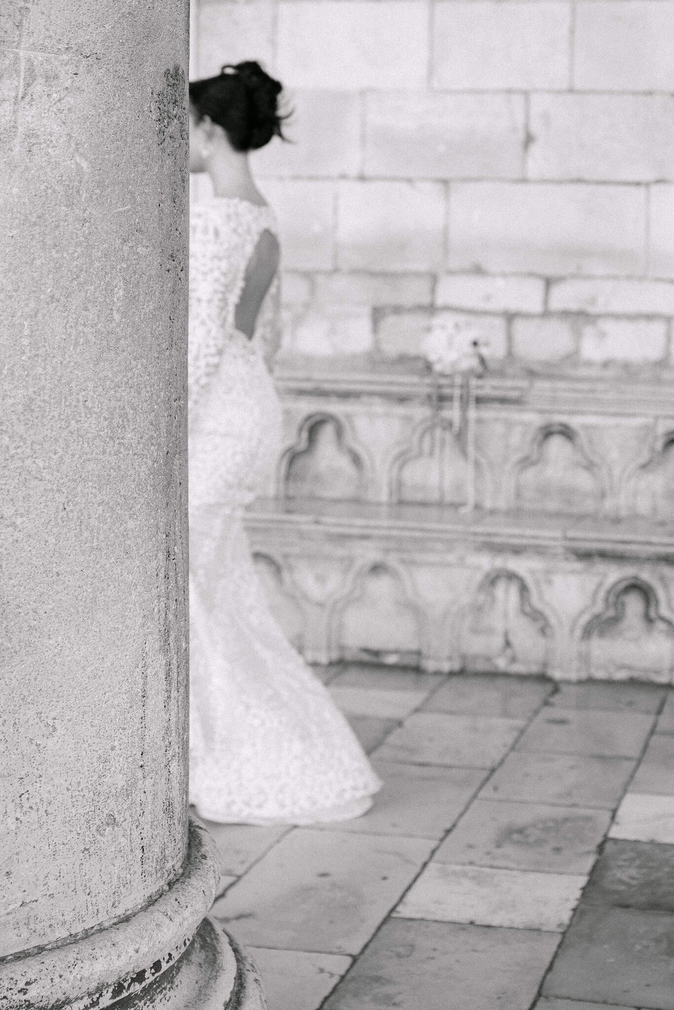 Vjenčanje u Dubrovniku na otvorenom – mladenka u čipkastoj vjenčanici s otvorenim leđima stoji iza povijesnog kamenog stupa.