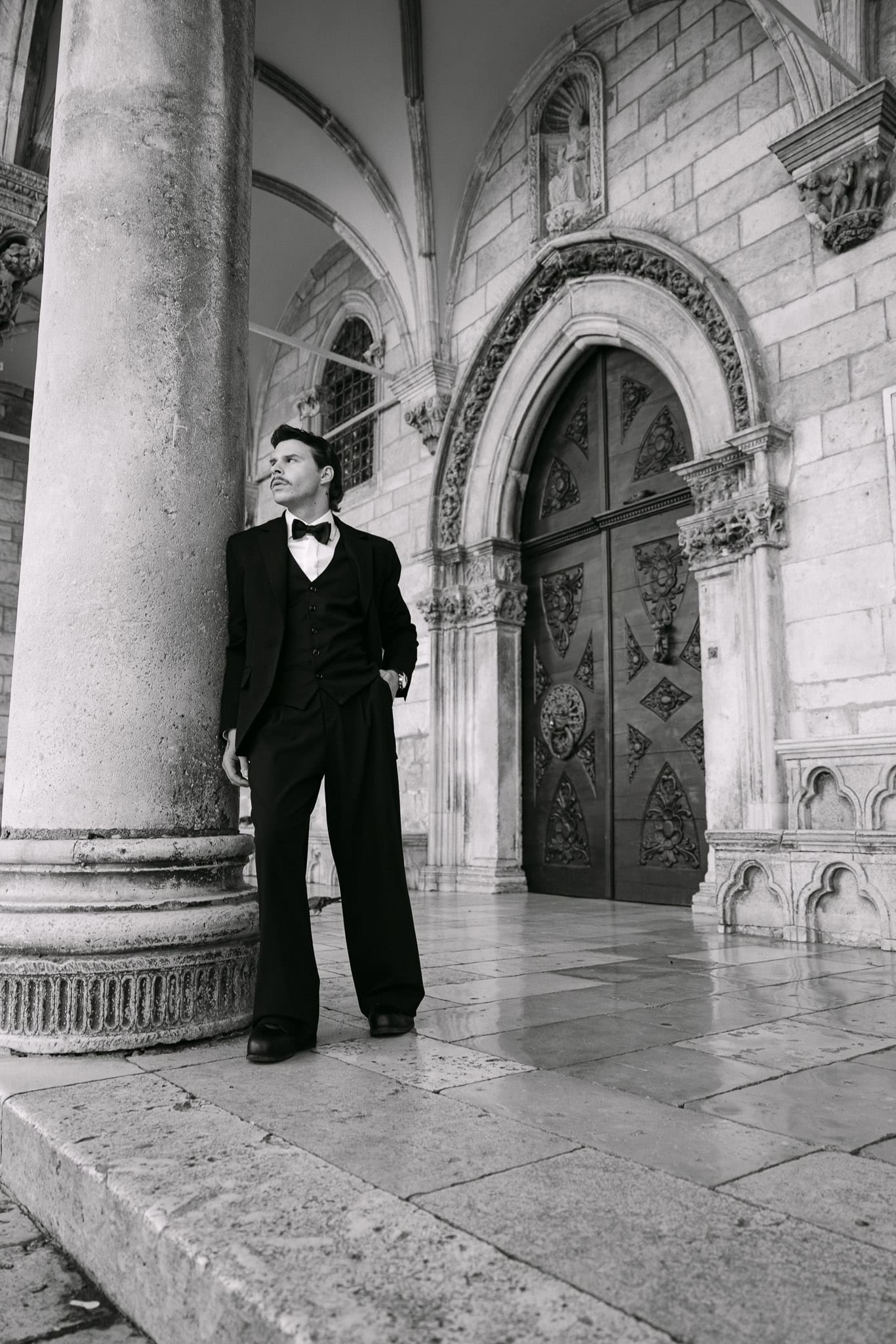 Vjenčani fotograf Dubrovnik – široki crno-bijeli kadar; mladoženja stoji uz kameni stup ispred impresivnih drvenih vrata s povijesnim dubrovačkim ornamentima.