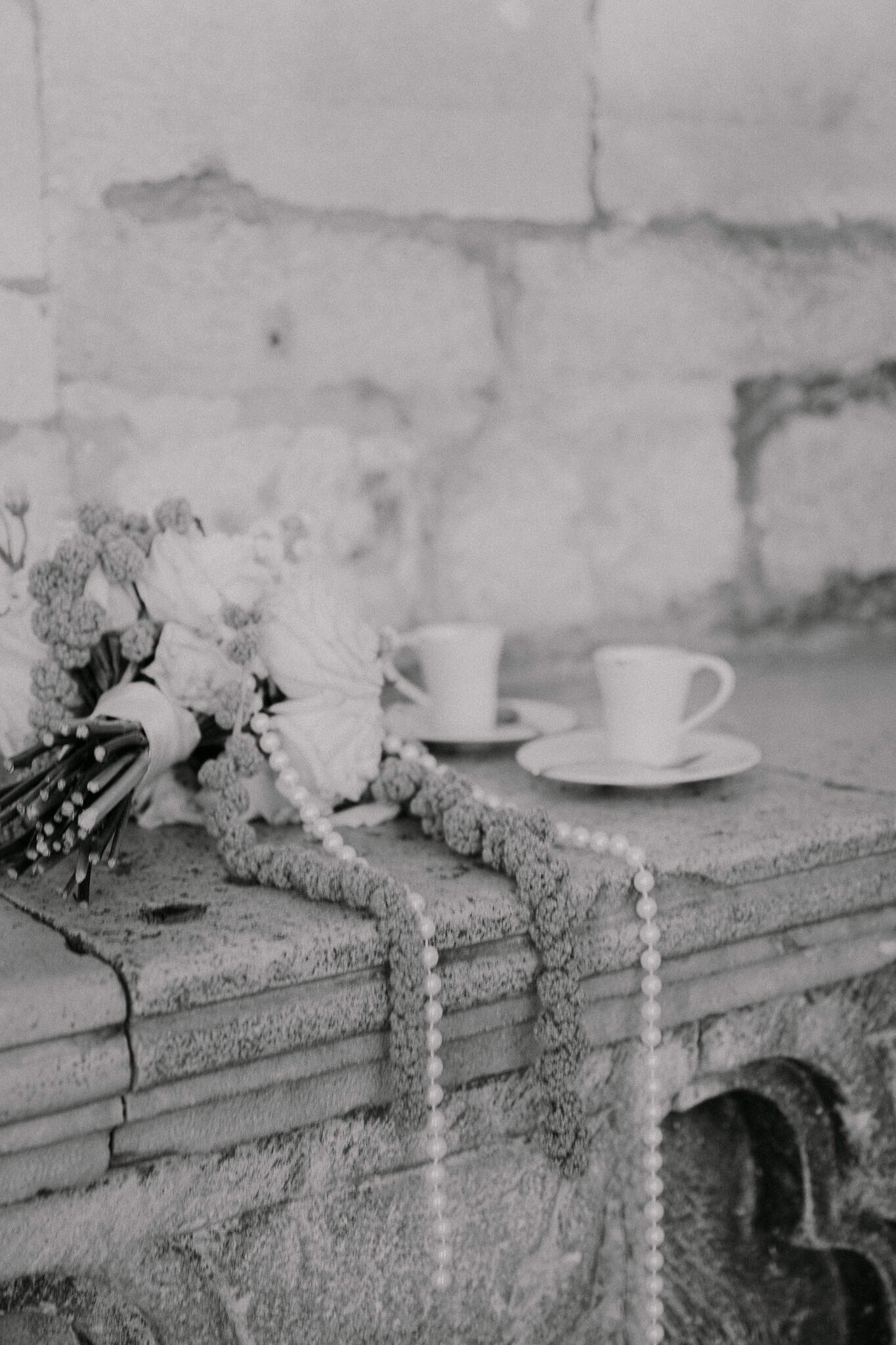 Vjenčani fotograf Dubrovnik – crno-bijela fotografija detalja; vjenčani bidermajer i šalice kave na staroj kamenoj klupi.