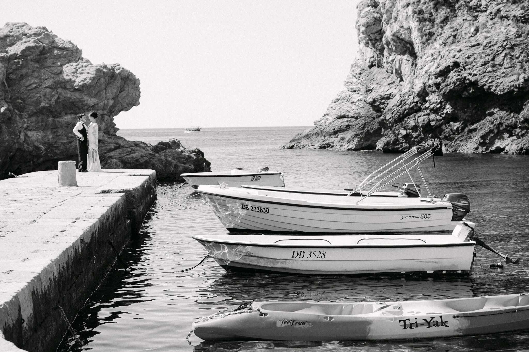Dubrovnik, morska obala - široki crno-bijeli kadar mladenaca; par stoji na starom kamenom mulu okružen drvenim brodicama i visokim stijenama.