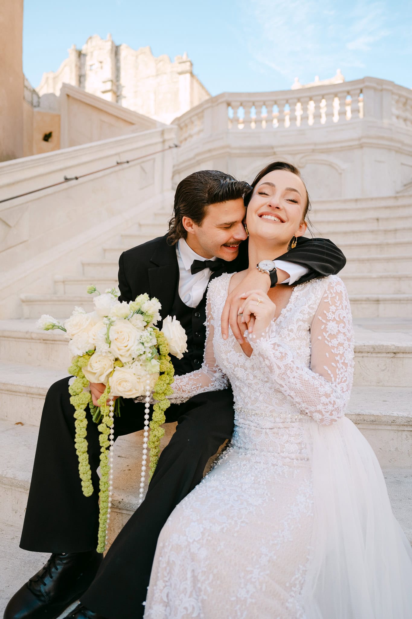 Radostan i opušten kadar na otvorenom; mladoženja grli nasmiješenu mladenku dok zajedno sjede na monumentalnom bijelom stubištu okruženi povijesnom arhitekturom grada Dubrovnika.