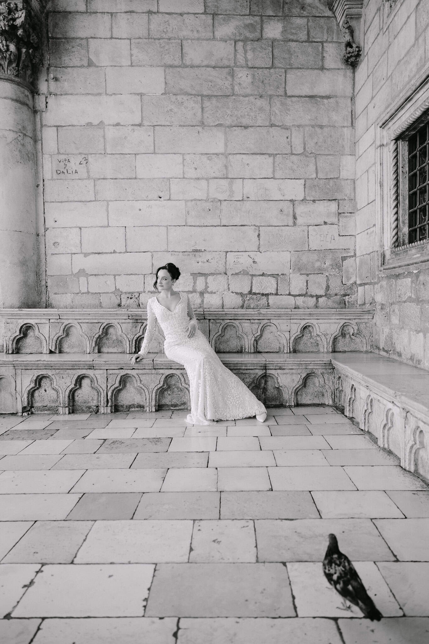 Vjenčanje Dubrovnik – široki crno-bijeli kadar arhitekture; mladenka pozira na kamenoj klupi unutar dubrovačkih zidina dok golub šeće u prvom planu.