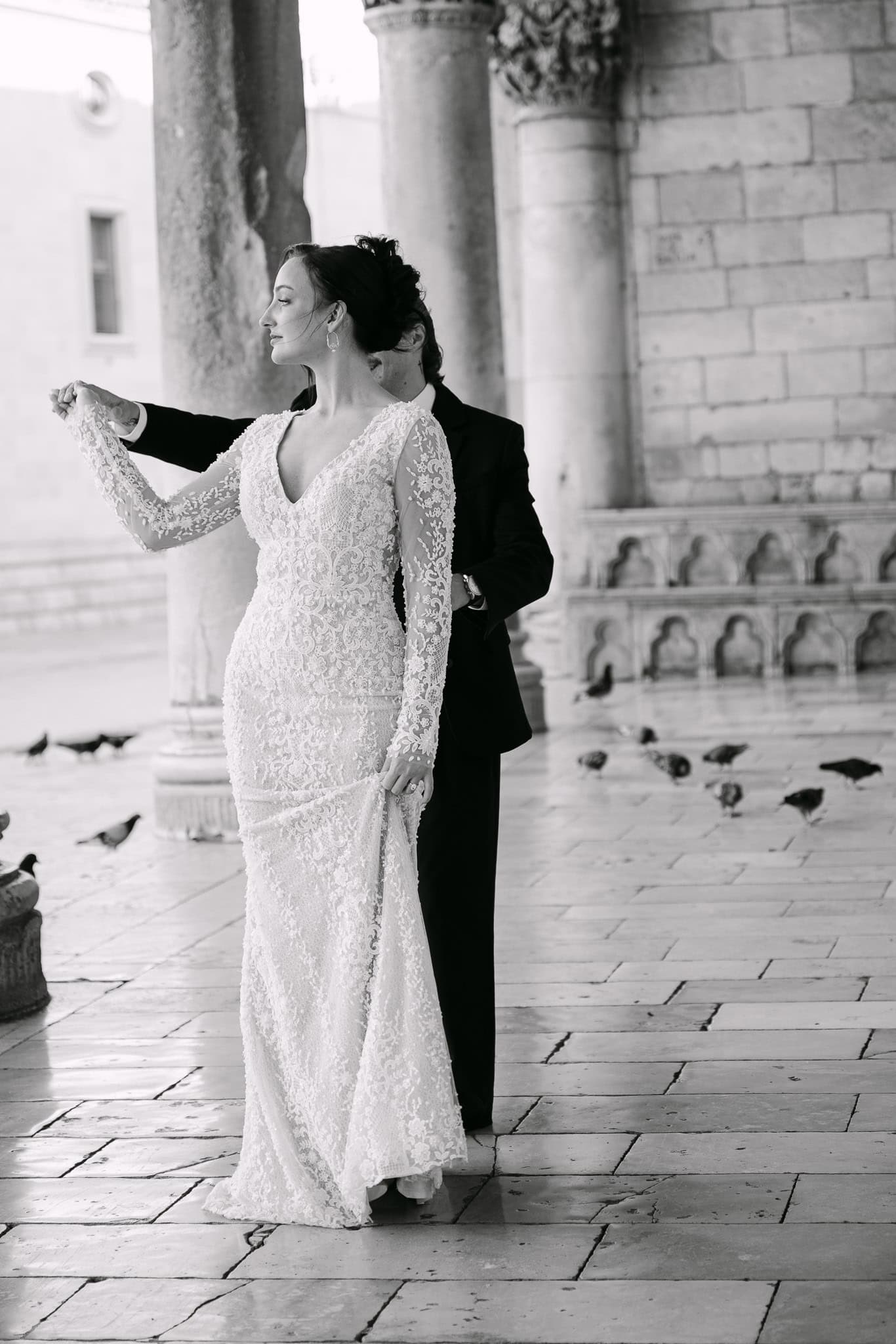 Vjenčani fotograf Dubrovnik – klasični crno-bijeli portret na otvorenom; mladoženja stoji iza mladenke držeći je za ruku ispred impresivnih povijesnih arkada, kamenih stupova i golubova u pozadini.
