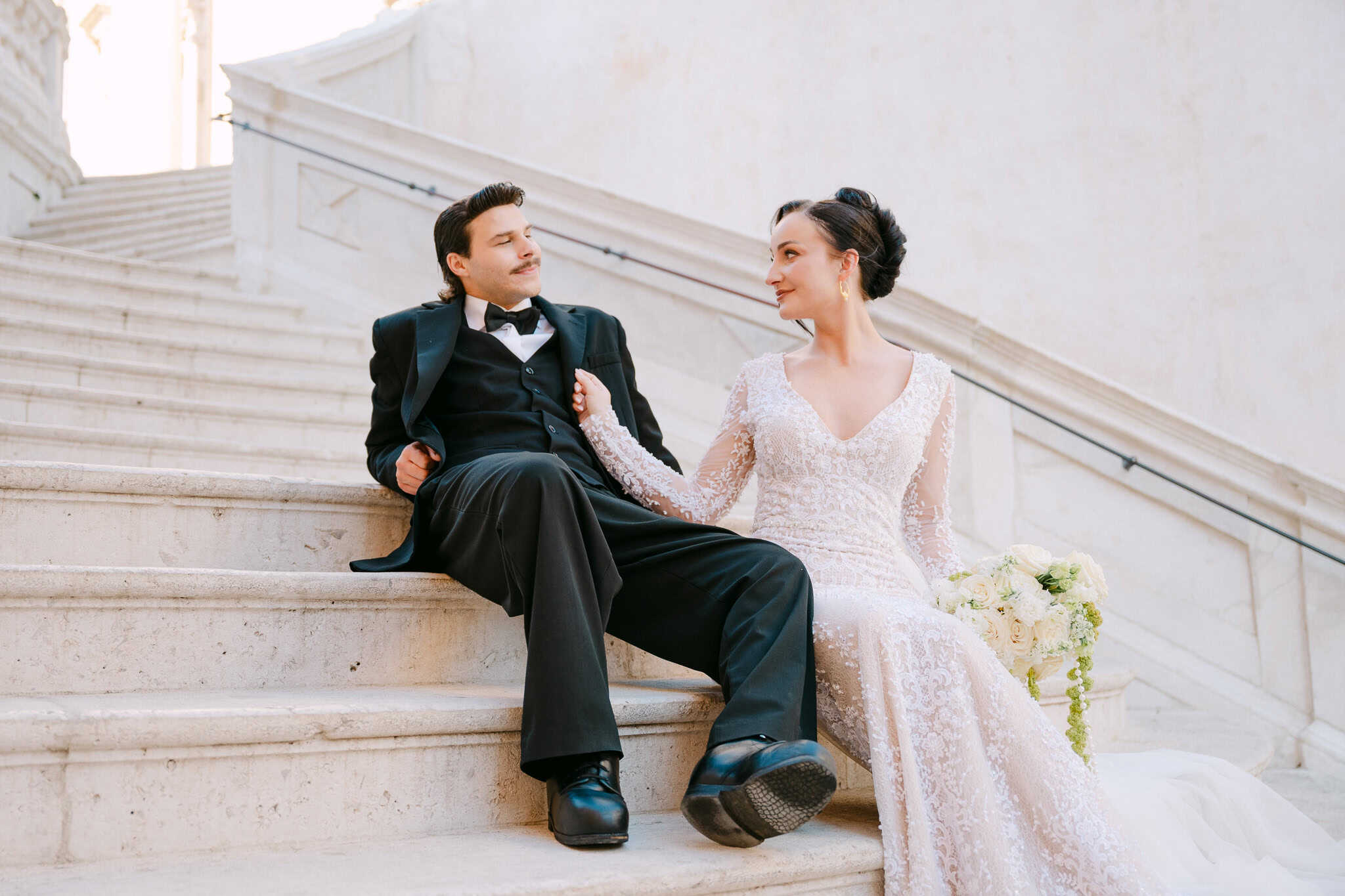 Topli lifestyle trenutak nakon ceremonije; par sjedi na bijelom kamenom stubištu i zaljubljeno se gleda, stvarajući prirodnu i nepoziranu atmosferu vjenčanog dana.