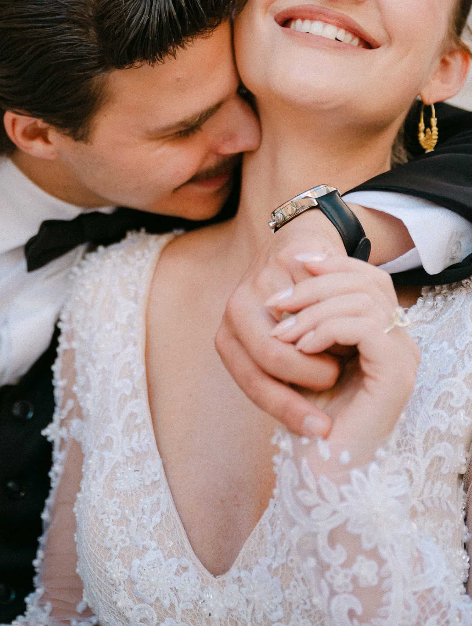 Romantični krupni plan pun emocija; mladoženja nježno ljubi mladenku u vrat, s jasnim fokusom na njene elegantne naušnice, teksturu čipke na haljini i njihove isprepletene ruke.