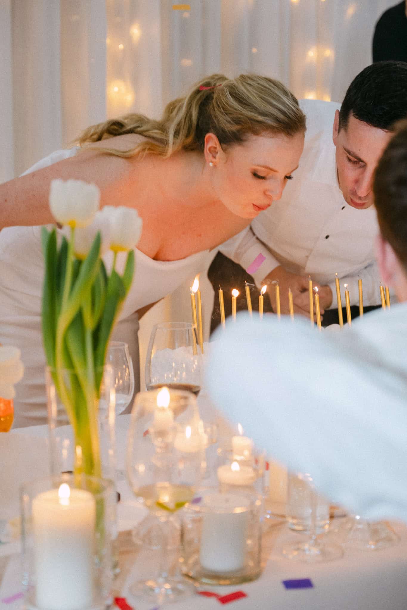 Topli krupni plan mladenaca tijekom svadbene torte; nevjesta i mladoženja posvećeni su tradiciji gašenja tankih svijeća na elegantno dekoriranom stolu s bijelim tulipanima.