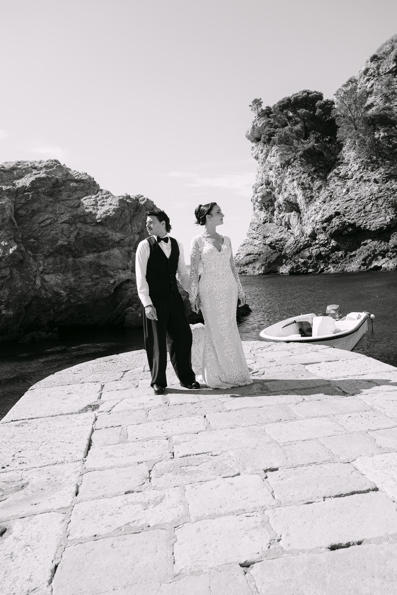 Crno-bijela vjenčana fotografija u modnom stilu; mladenci poziraju okrenuti u suprotnim smjerovima na kamenom pristaništu u Dubrovniku ispred Lovrijenca.
