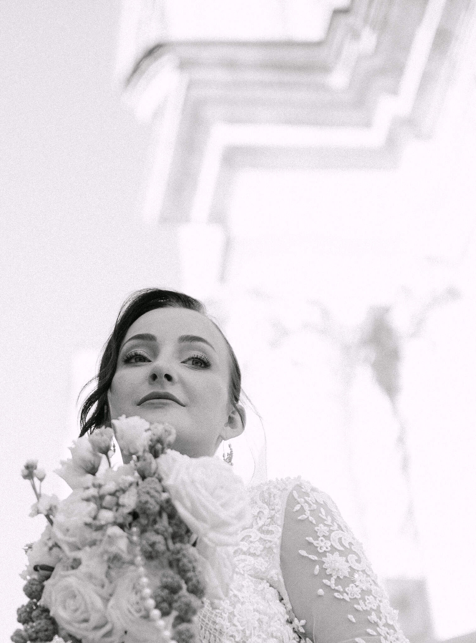 Dramatičan crno-bijeli portret mladenke snimljen iz donjeg kuta; nevjesta s raskošnim cvjetnim bidermajerom ispred grandiozne arhitekture Dubrovnika.