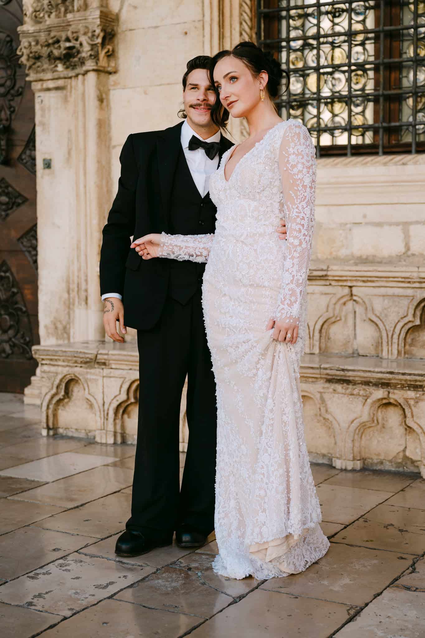 Vjenčanje u Dubrovniku na otvorenom – klasični portret mladenaca u punoj visini; mladoženja u crnom odijelu i nevjesta u dugoj čipkastoj vjenčanici poziraju ispred povijesne kamene arhitekture i masivnih drvenih vrata Starog grada.