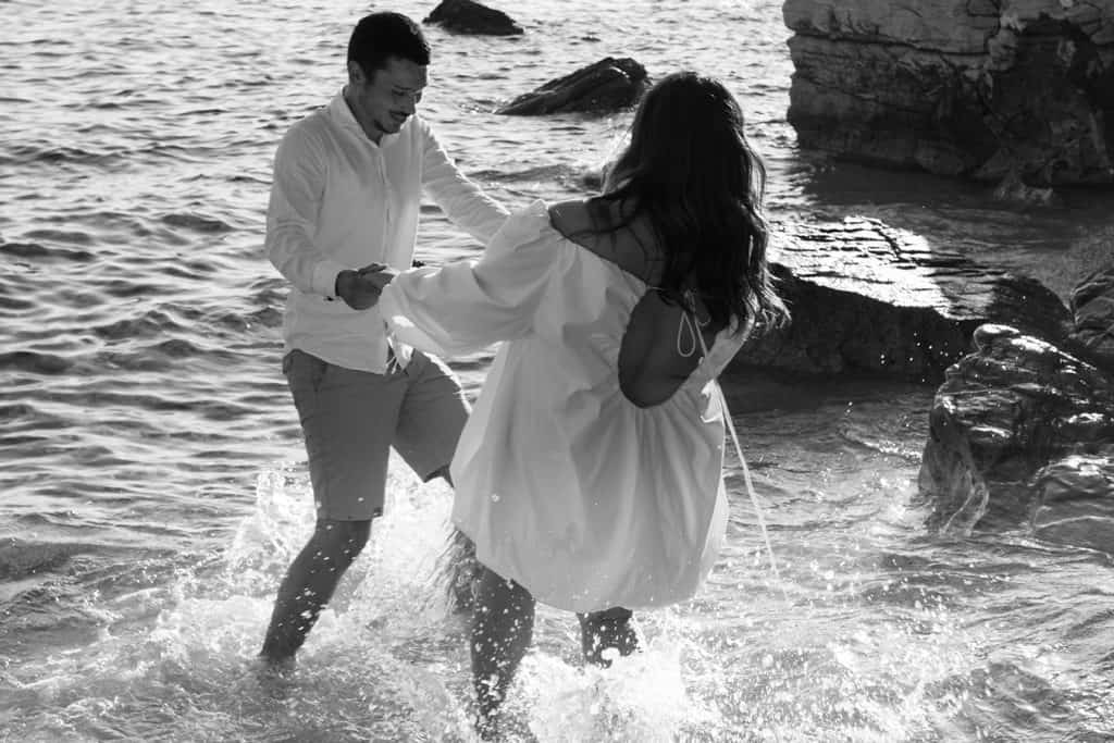 Crno-bijela fotografija zaručničkog para koji se drži za ruke i gazi kroz plićak mora tijekom fotografiranja na plaži.